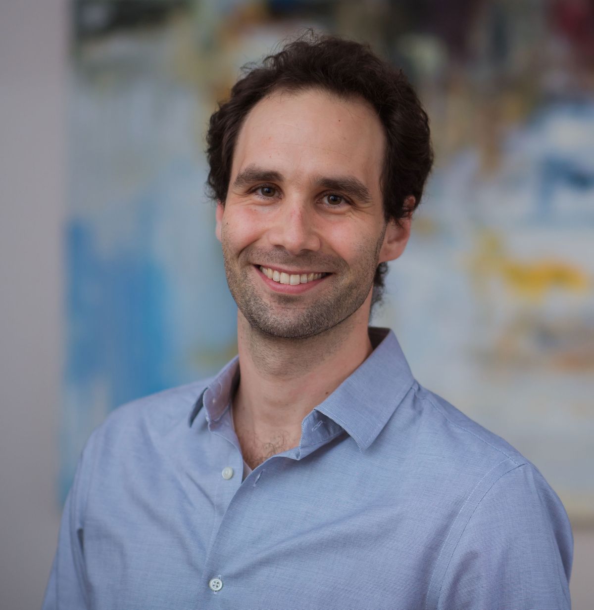 The researcher Siniša Hrvatin wears a blue shirt and smiles at the camera. He stands in front of a painting. The researcher Siniša Hrvatin wears a blue shirt and smiles at the camera. He stands in front of a painting.