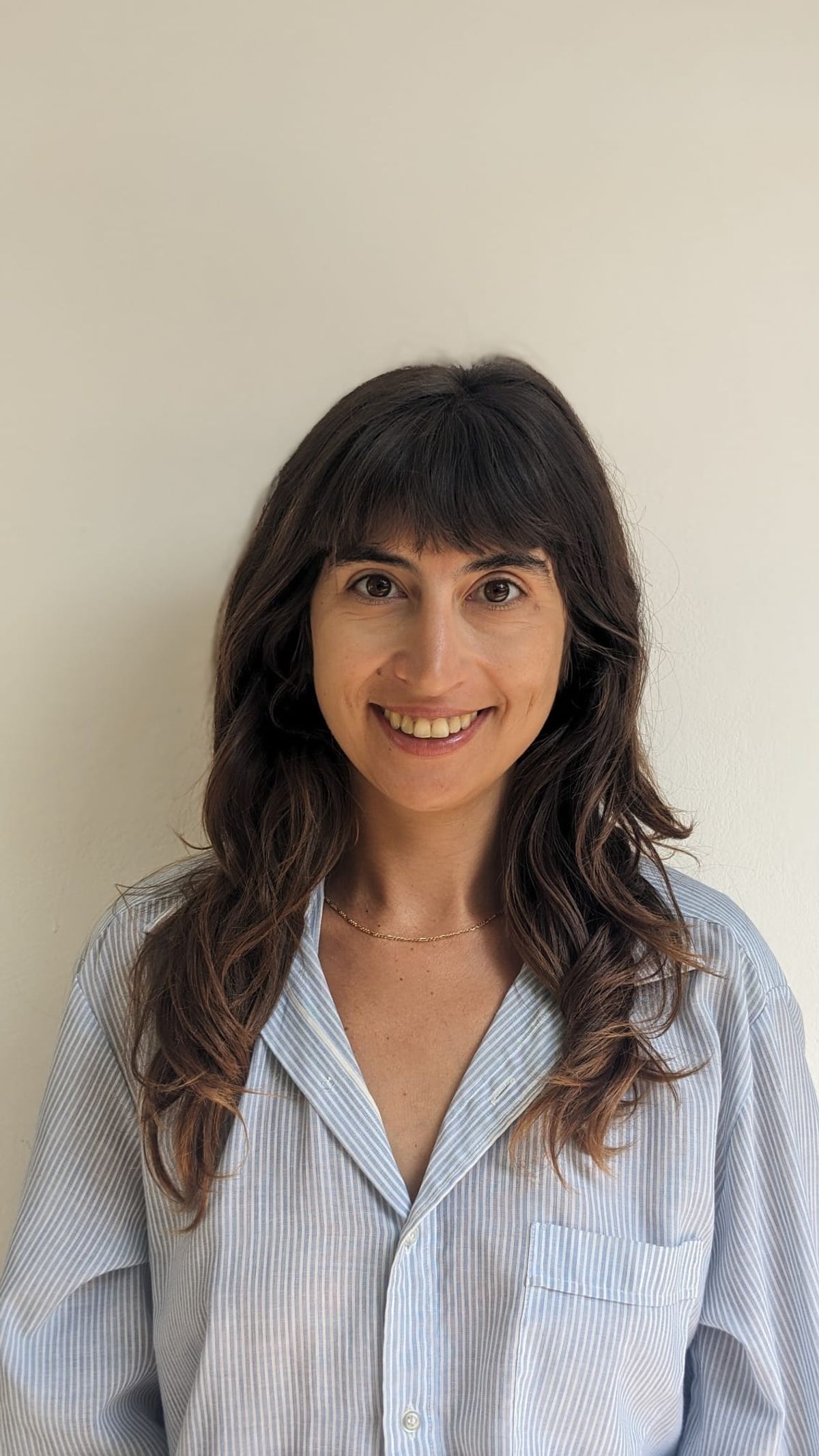 Hedva Haykin wears a blue button-down shirt and smiles in front of a white wall. Hedva Haykin wears a blue button-down shirt and smiles in front of a white wall.