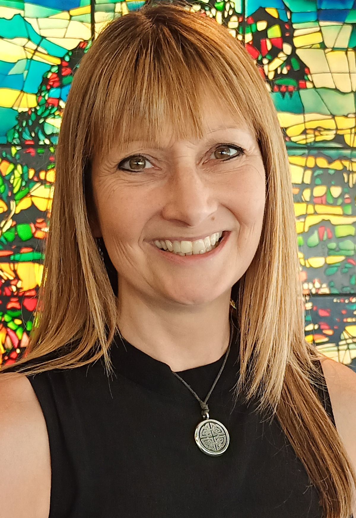 A woman wears a black sleeveless shirt and smiles at the camera. She stands in front of a stained-glass window. A woman wears a black sleeveless shirt and smiles at the camera. She stands in front of a stained-glass window.