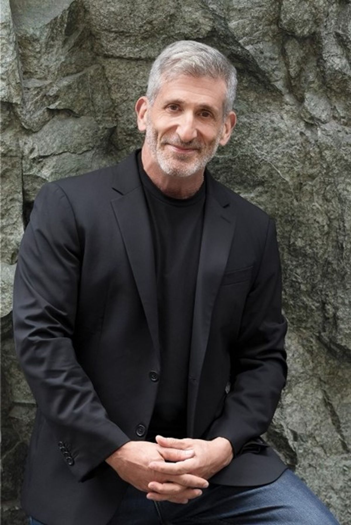 Photo of a scientist with interlaced fingers, wearing a black shirt and blazer, leaning against a rocky backdrop outdoors. Photo of a scientist with interlaced fingers, wearing a black shirt and blazer, leaning against a rocky backdrop outdoors.
