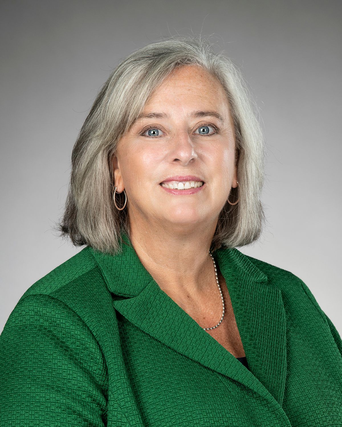 The photo shows a woman wearing a green blazer and smiling at the camera. The photo shows a woman wearing a green blazer and smiling at the camera.