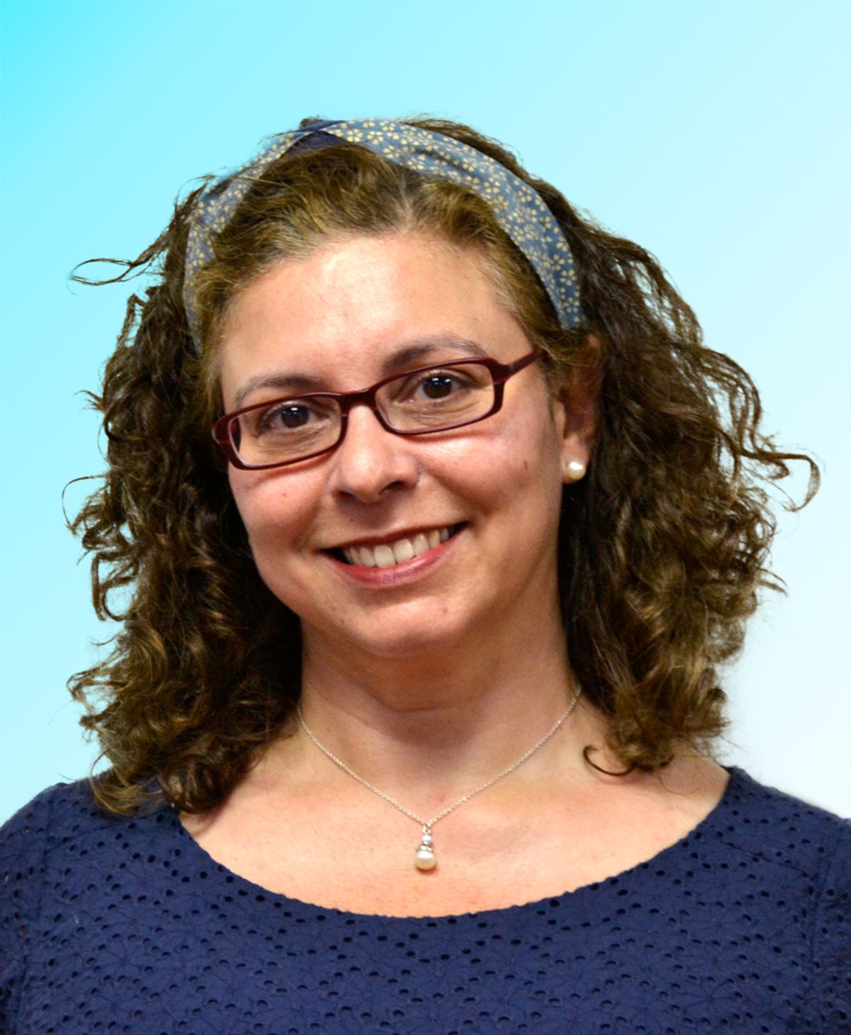 A woman with wavy hair wears a blue shirt and glasses. She smiles at the camera. A woman with wavy hair wears a blue shirt and glasses. She smiles at the camera.